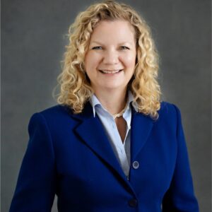 caucasian female with curly blonde hair wearing a blue jacket and colored shirt with a grey background