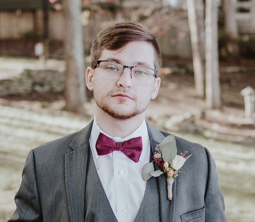 Caucasian Man with beard and glasses wearing a grey suit and black bowtie with a Boutonniere.