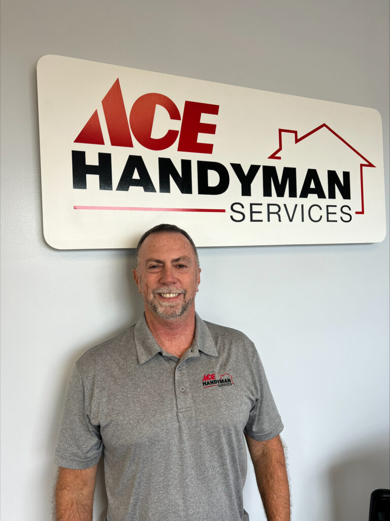 Older man smiling in a grey polo shirt, in front of a sign that says "Ace Handyman Services"